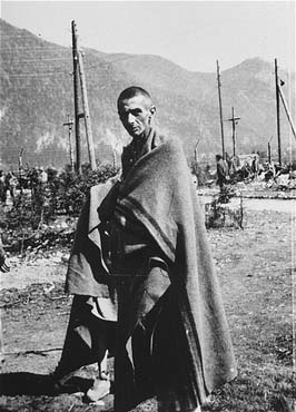an emaciated survivor stands outside wrapped in a blanket in the Ebensee concentration camp.jpg