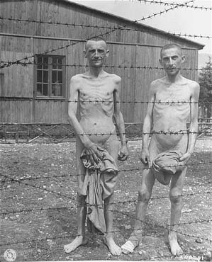 Two emaciated survivors stand behind a barbed wire fence in the hospital compound at the Ebensee.jpg