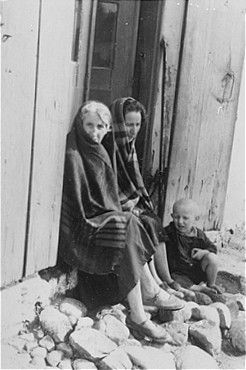 Two Jewish women and a child sit in the doorway of a wooden house in Konskowola.jpg