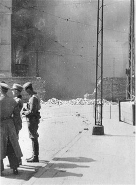German personnel discuss matters as an apartment building razed by the SS burns.jpg